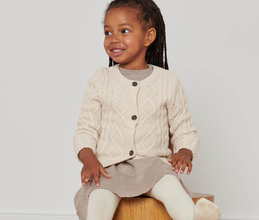 Lächelndes Mädchen sitzt auf einem Holzhocker und trägt eine Strickjacke mit RWS-Wolle.