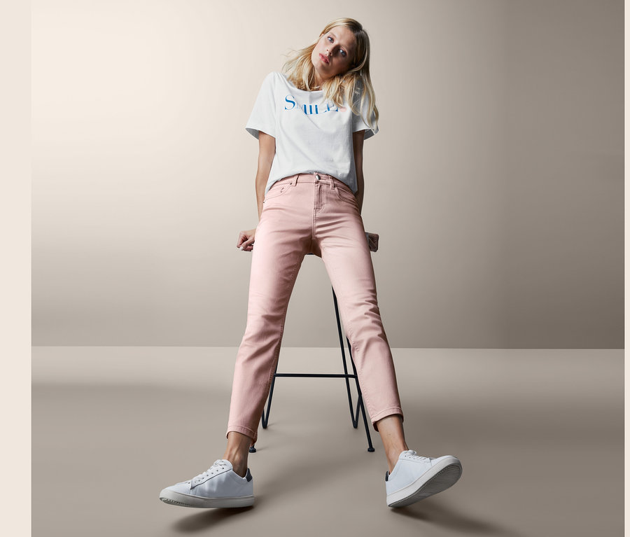 Blonde Frau sitzt auf einem Hocker und trägt eine rosafarbene Slimfit-Jeans und weiße Turnschuhe.
