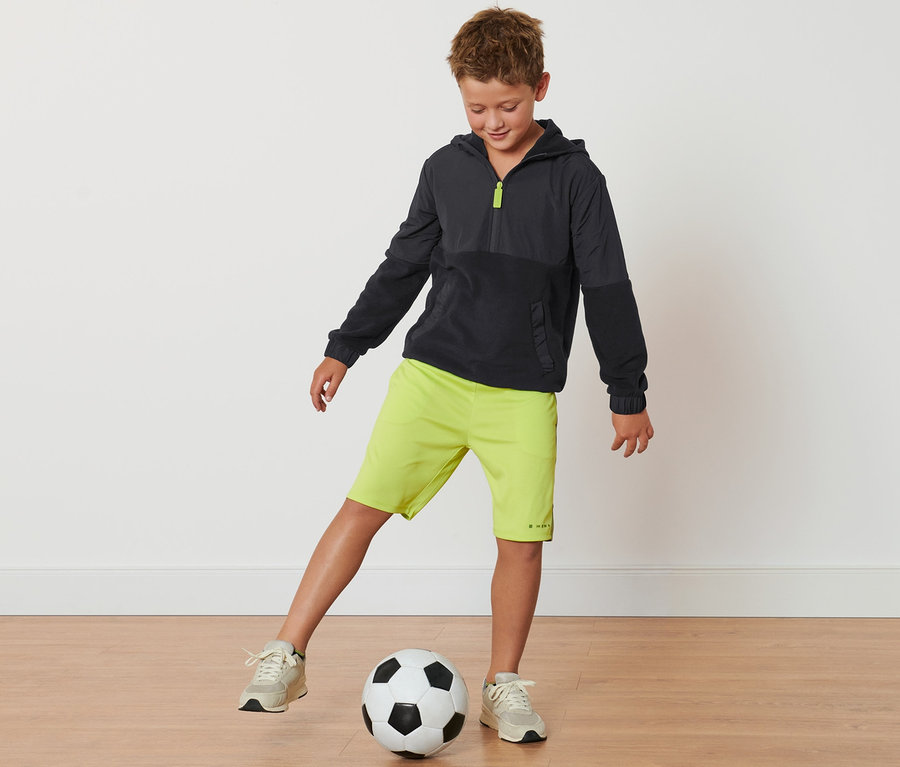 Ein Junge tritt gegen einen Fußball. Er trägt einen Kinder-Fleecesweater, neon-grüne Shorts und Turnschuhe.