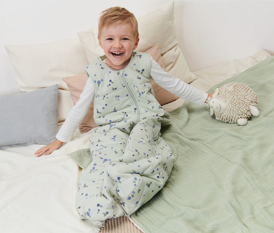 Ein lächelnder Junge sitzt in einem Schlafsack 2,5 TOG, 110 cm, Beeren auf einem Bett und hält ein Plüschtier.