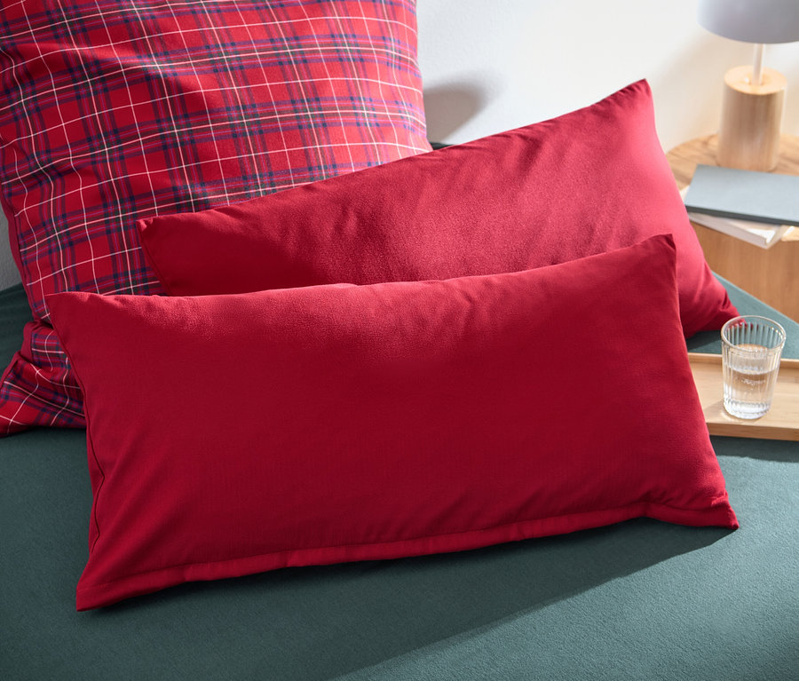 Auf einem Bett liegen zwei rote Microfaser-Flanell-Kopfkissenbezüge, ca. 80 x 40 cm und ein karierter Kissenbezug.