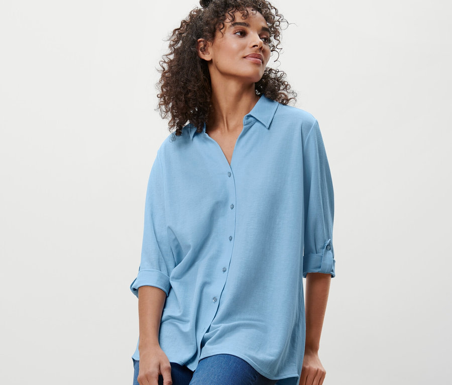 Eine Frau sitzt auf einem Hocker und trägt eine hellblaue Piqué-Jerseybluse und Jeans.