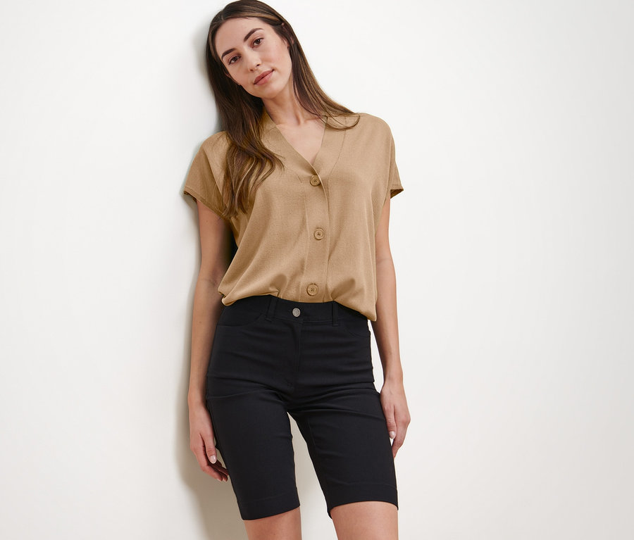Frau posiert in beiger Bluse und schwarzen Bengalin-Bermudashorts.