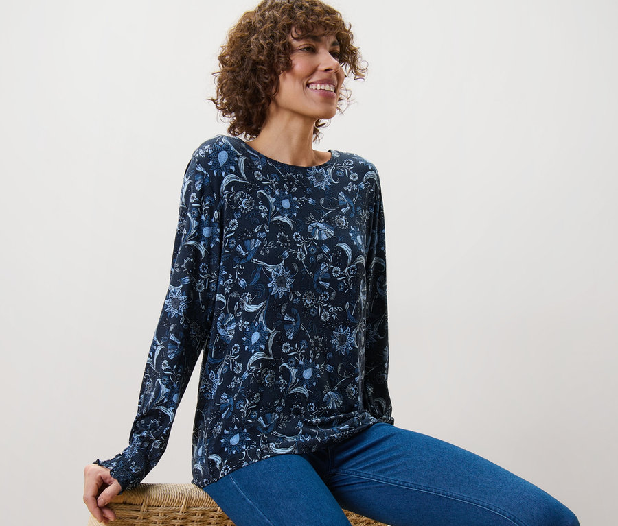 Frau sitzt auf einem Korbstuhl und trägt ein bedrucktes Blusenshirt.