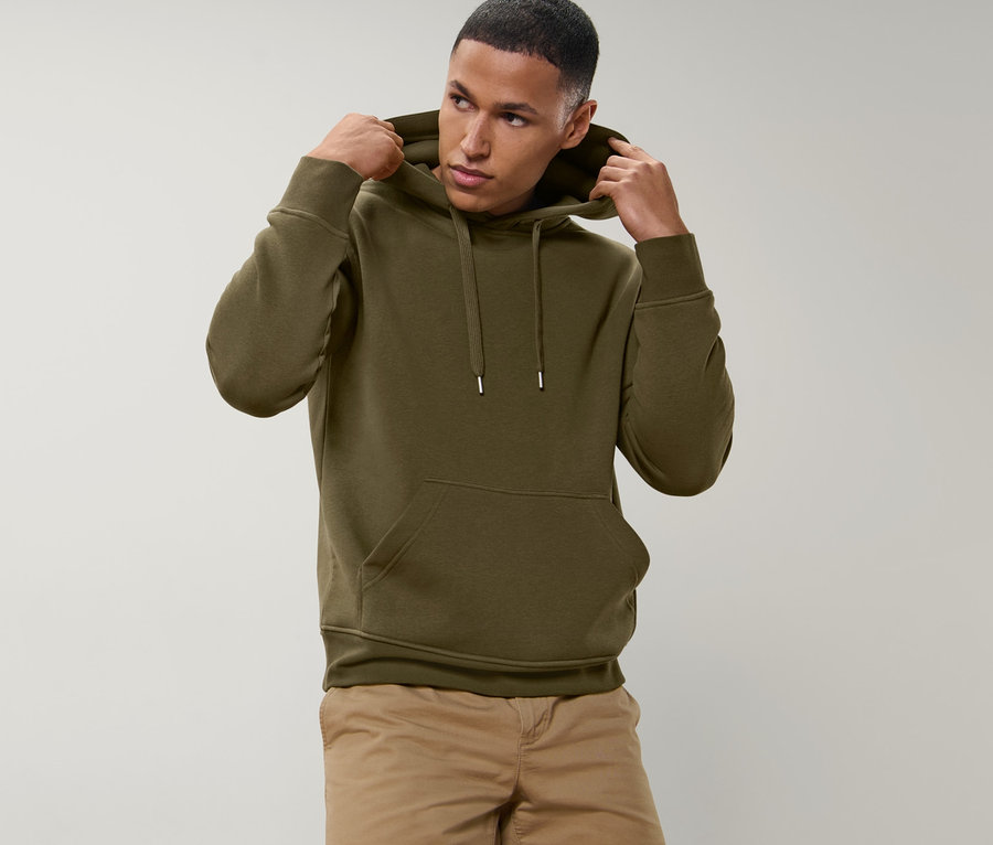 Mann richtet die Kapuze eines olivgrünen Hoodies. Er trägt beige Shorts.