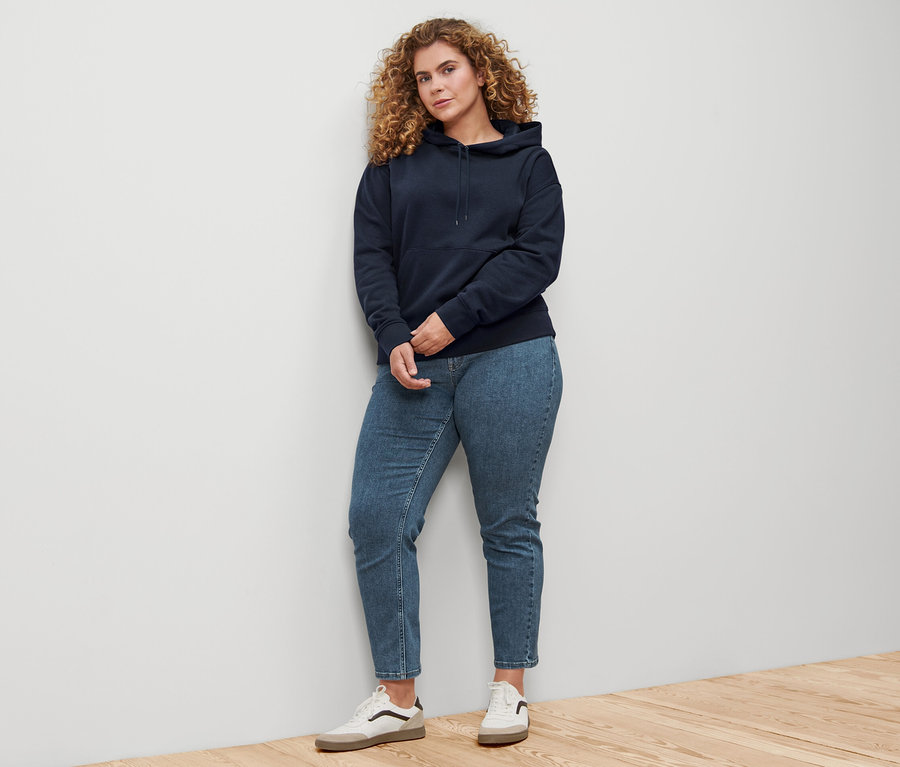 Frau steht an einer Wand und trägt einen dunkelblauen Hoodie.