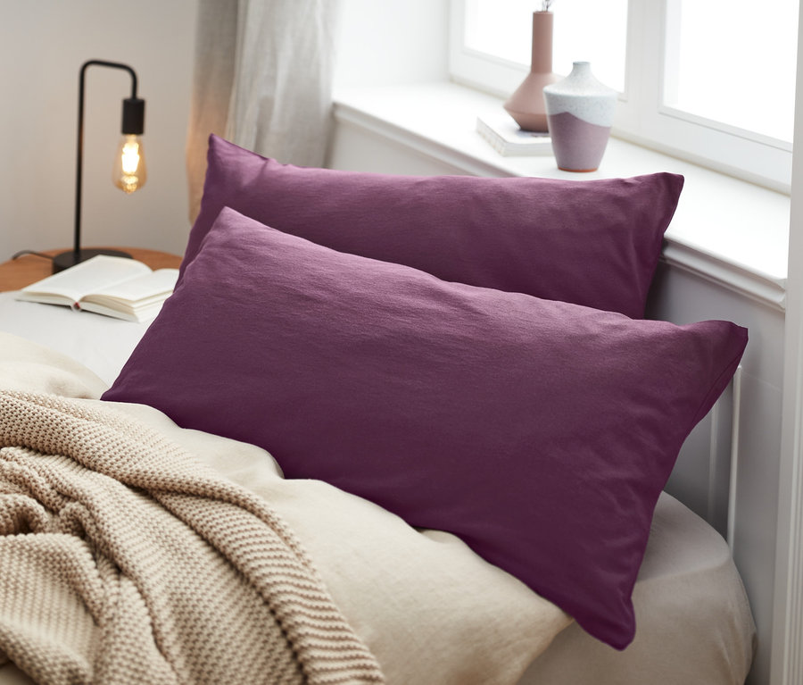 Zwei lila Jersey-Kopfkissenbezüge liegen auf einem Bett mit einer beige Decke.
