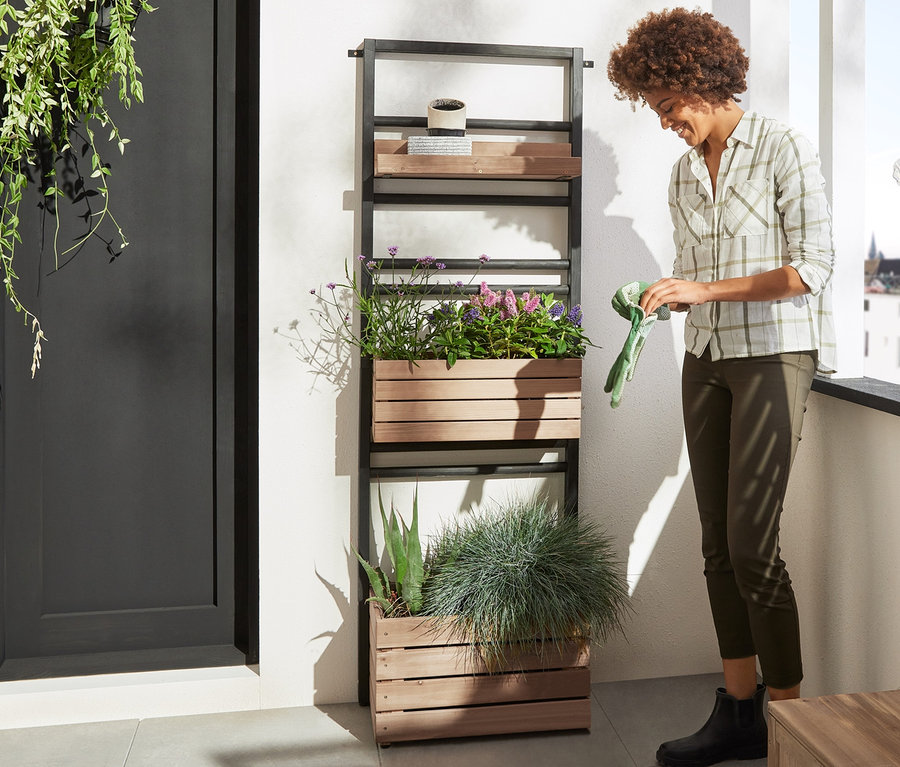 Frau zieht Gartenhandschuhe neben Pflanzwand »Elin« mit flexibel einhängbaren Regalen und Sichtschutz-Seiten-Modul »Elin« mit Stauraum-Bank an.