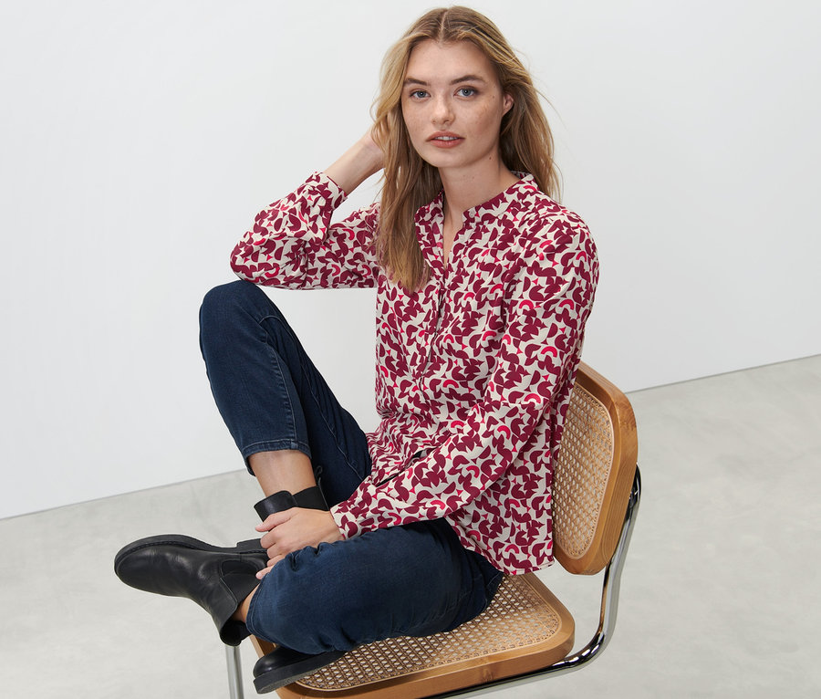 Frau sitzt auf einem Stuhl und trägt eine bedruckte Bluse.