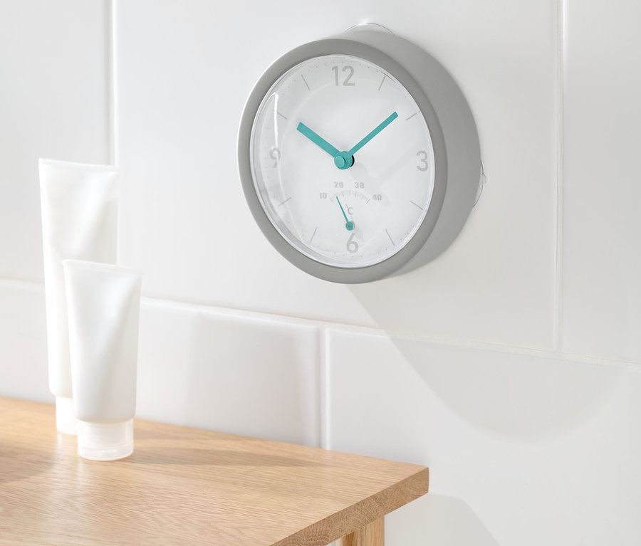 Eine Funk-Badwanduhr mit Thermometer hängt an der weißen Badezimmerwand. Zwei weiße Tuben stehen auf einem Holztisch.