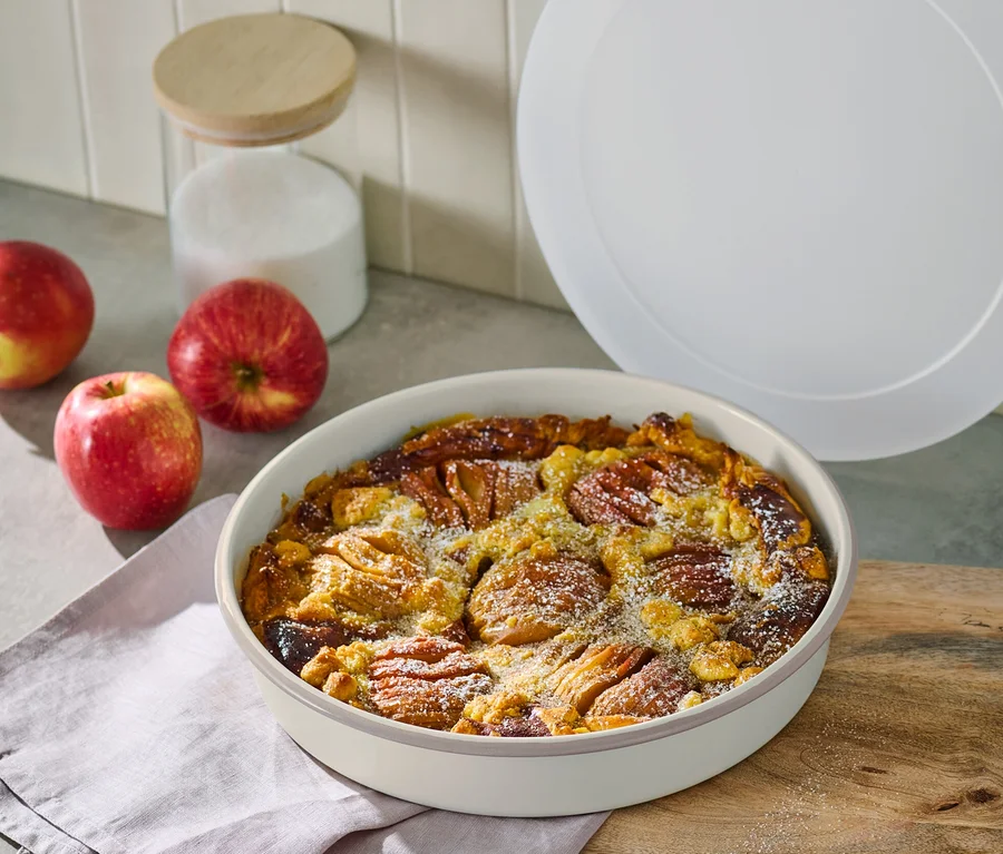 Apfelkuchen in Emaille-Tarteform auf Holzbrett neben Äpfeln und Zuckerdose.