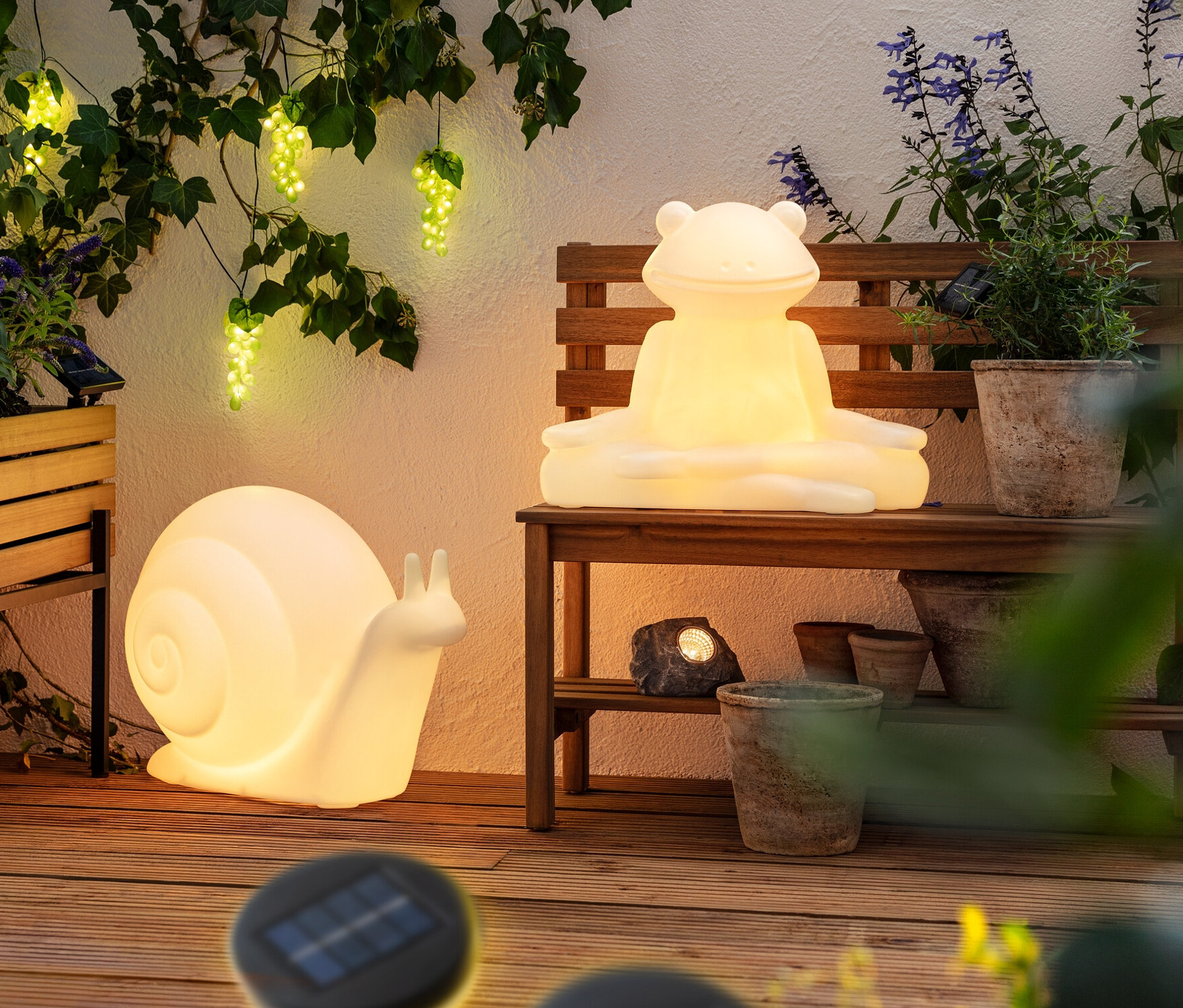 Auf einer Terrasse sind ein Pflanzkasten »Elin«, eine Solar-Lichterkette »Weintrauben«, eine Solar-Leuchte »Frosch«, eine Solar-Leuchte »Schnecke« und 3 Solar-LED-Steine zu sehen.