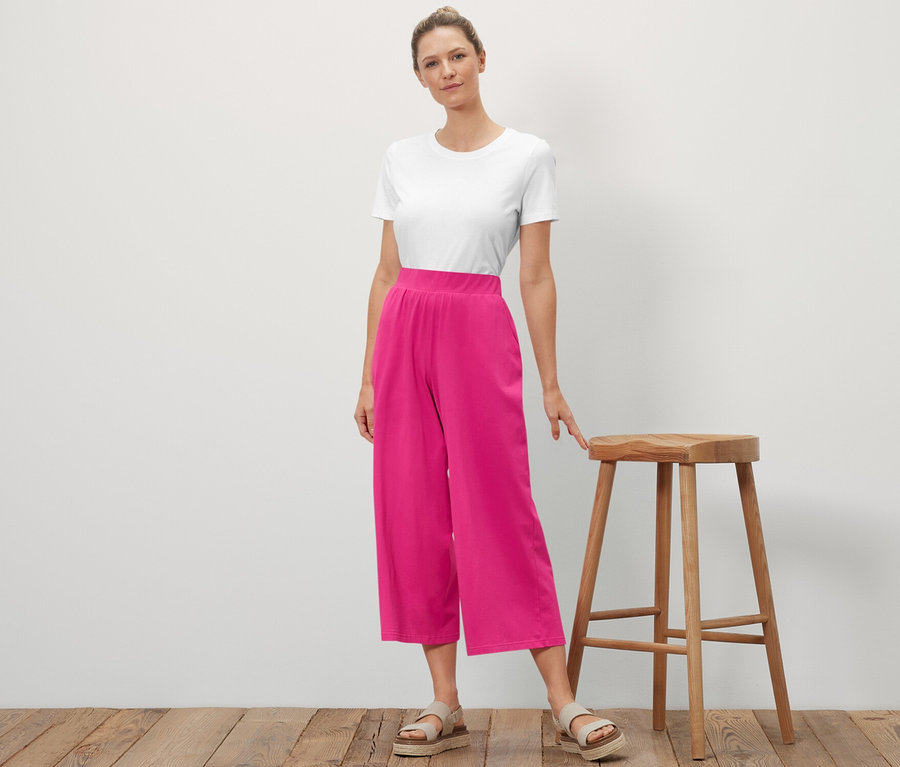 Eine Frau steht neben einem Holzhocker und posiert. Sie trägt ein weißes T-Shirt und eine rosafarbene Jersey-Culotte.