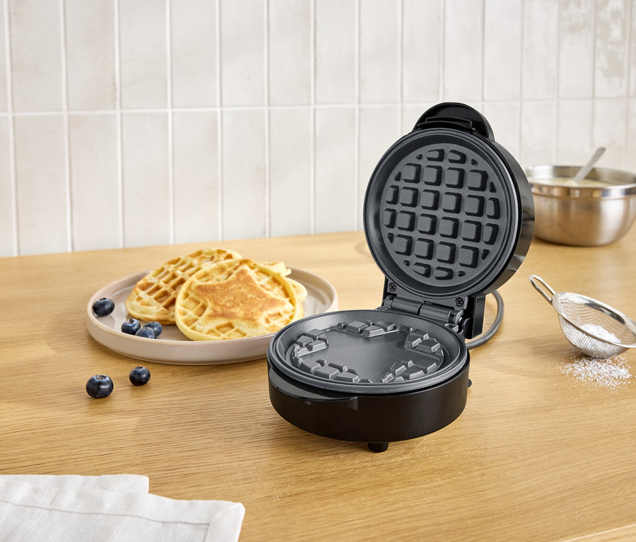 Offener, schwarzer Waffelautomat neben einem Teller mit Waffeln und Blaubeeren auf einem Holztisch.