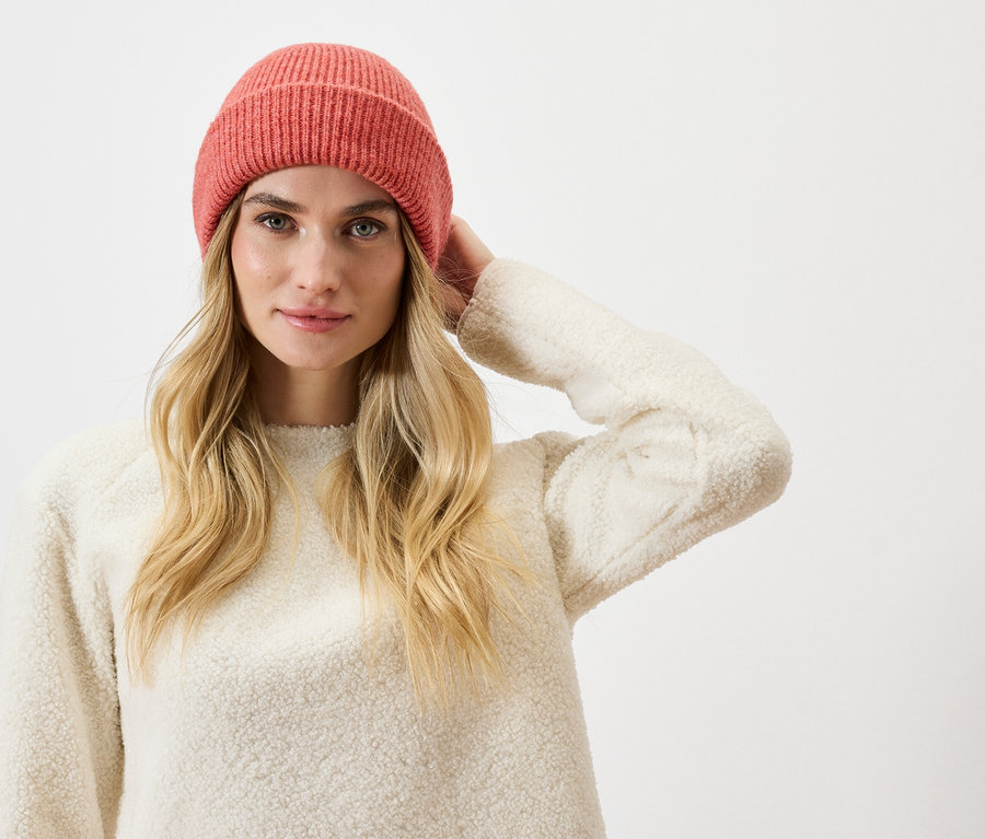 Frau mit blonden Haaren, einer rosafarbenen Strickmütze und einem cremefarbenen Pullover.
