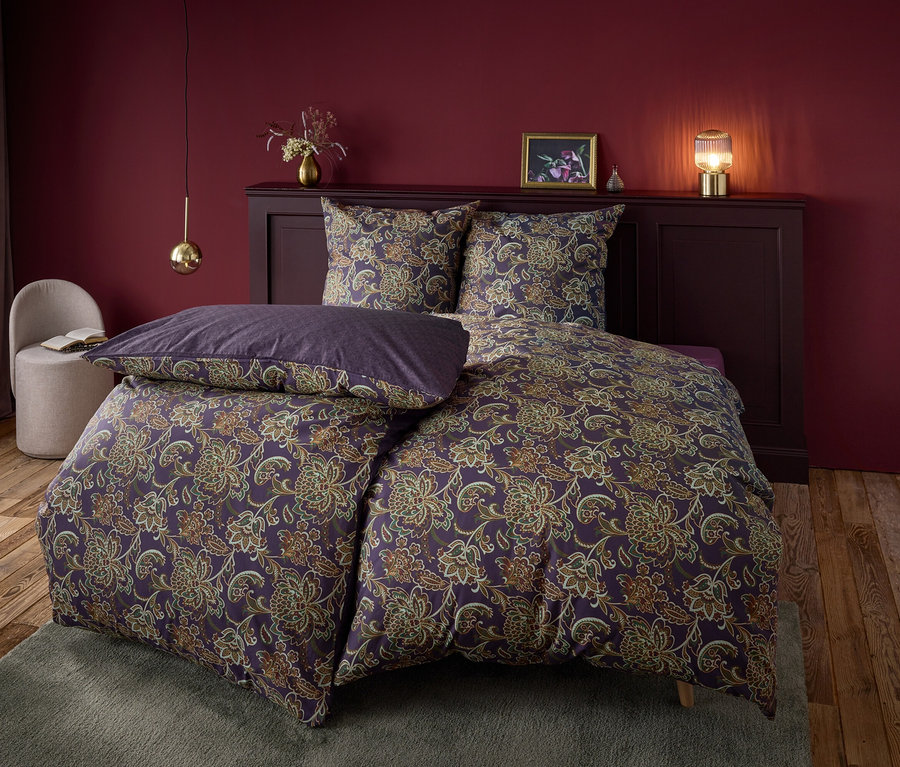 Lila-farbene Bettwäsche mit Blumenmuster auf einem Bett mit lila Kopfteil und Nachttisch vor einem dunkelroten Hintergrund.