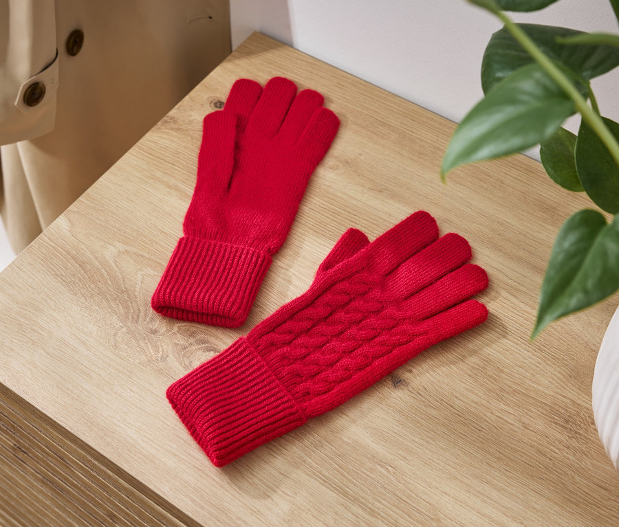 Zwei rote Strickhandschuhe liegen auf einem Holztisch neben einer Pflanze in einem weißen Topf.