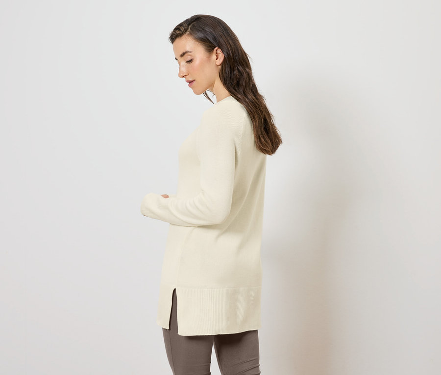 Frau posiert seitlich in einem langen Strickpullover in Creme.