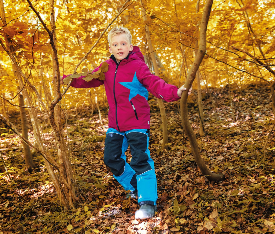 Ein Junge in einer pinkfarbenen Jacke mit blauem Stern und blau-schwarzen Hosen geht durch einen Herbstwald.
