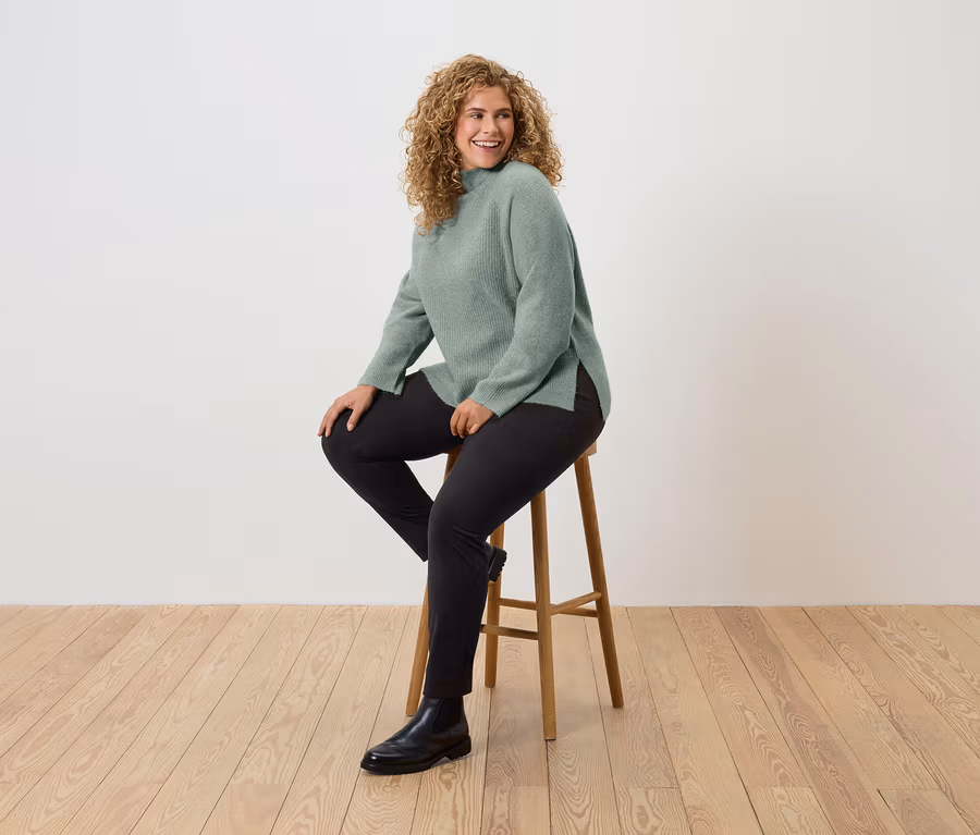 Eine Frau mit lockigem Haar sitzt auf einem Holzhocker und trägt einen grünen Pullover, schwarze Hosen und schwarze Stiefel.