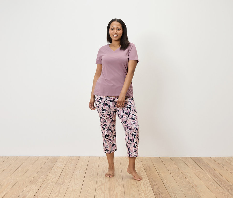 Eine Frau steht und trägt ein lila T-Shirt und eine geblümte Hose.