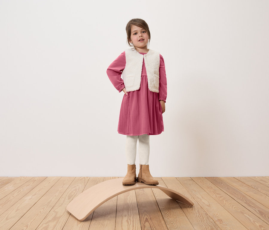 Mädchen steht auf einem hölzernen Balance Board, trägt ein rosa Kleid, eine weiße Weste und beige Stiefel.