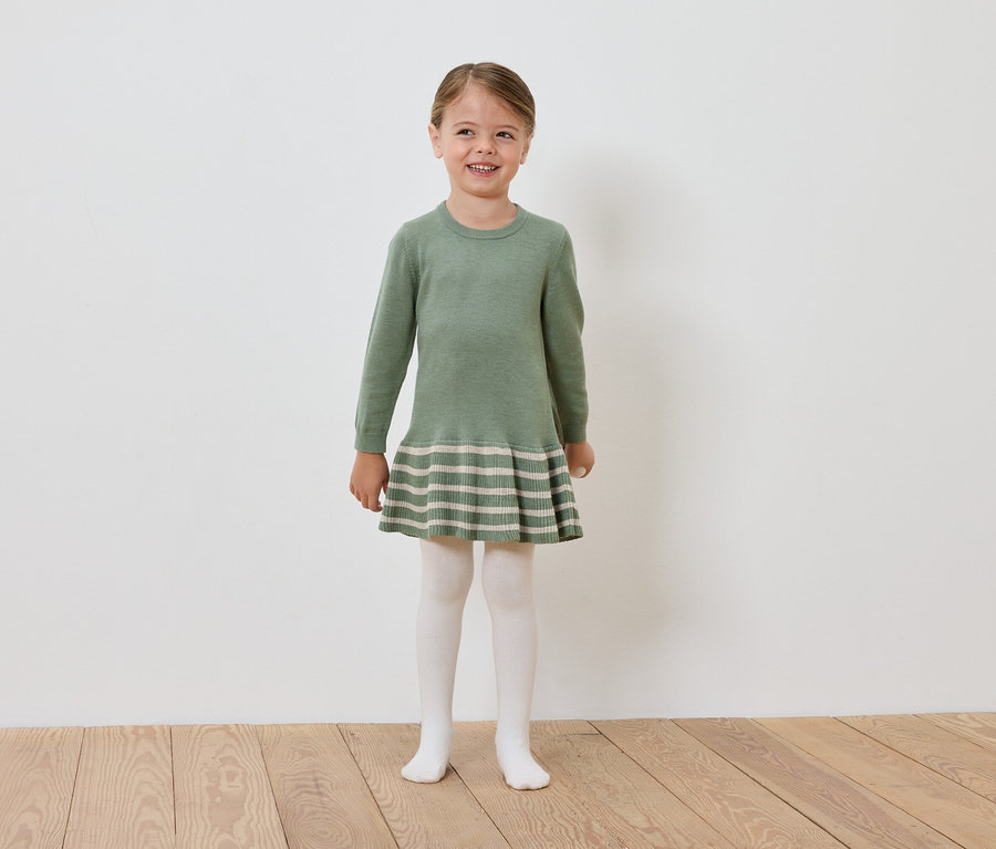 Ein lächelndes Mädchen in einem grünen Strickkleid und einer weißen Strumpfhose steht auf einem Holzboden vor einer weißen Wand.