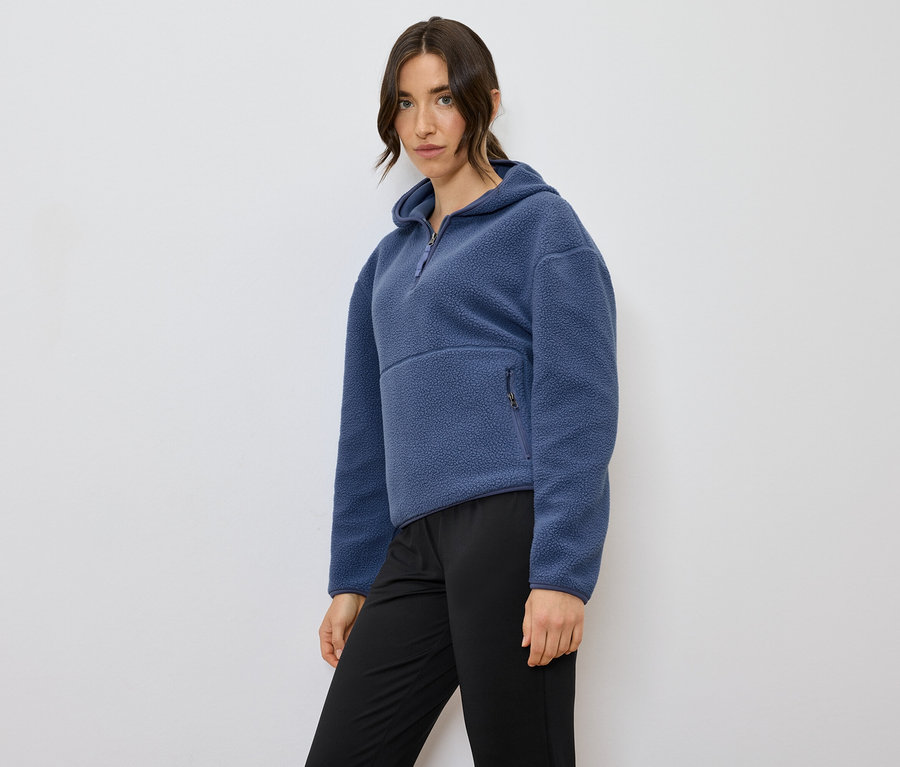 Frau posiert in einem blauen Fleecepullover mit Kapuze und schwarzen Hosen.
