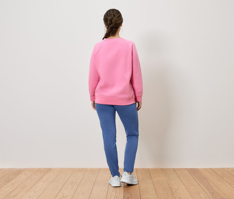 Eine Frau mit dem Rücken zur Kamera trägt ein rosa Sweatshirt, blaue Hosen und weiße Schuhe.