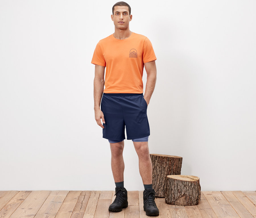 Mann posiert in 2-in-1-Funktionsshorts, orangefarbenem T-Shirt und Wanderstiefeln.