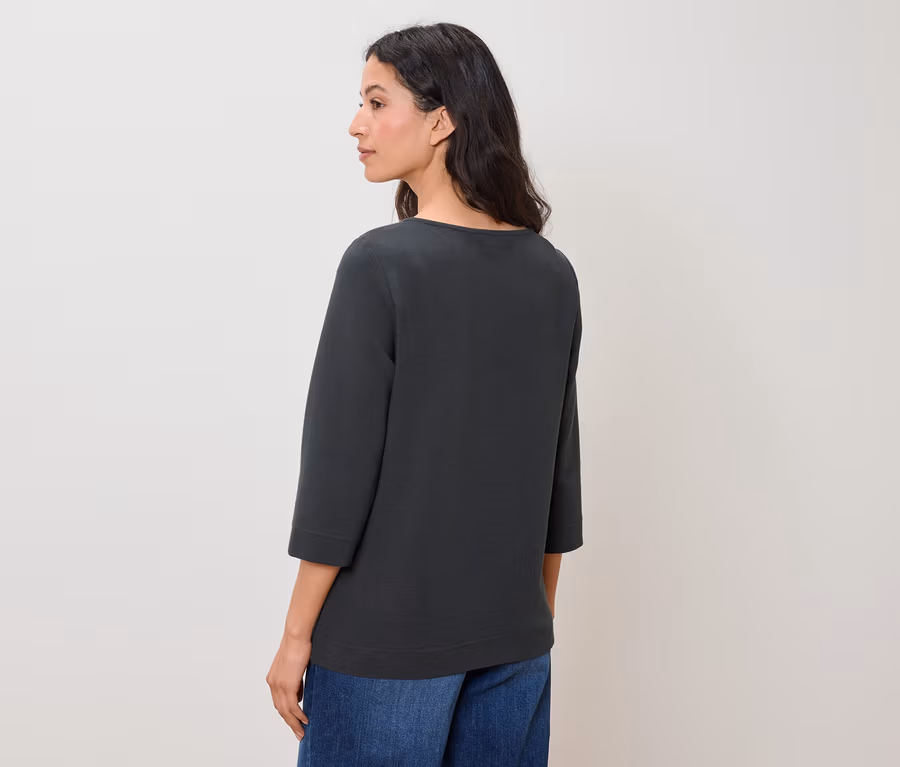 Eine Frau schaut nach links und trägt blaue Jeans und ein graugrünes Oberteil mit Dreiviertelärmeln.