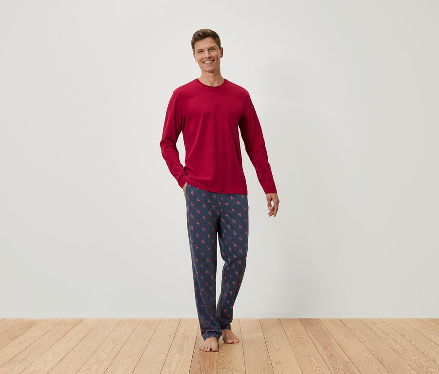 Mann steht barfuß, trägt ein rotes Langarmshirt und eine blaue Schlafanzughose mit Muster.