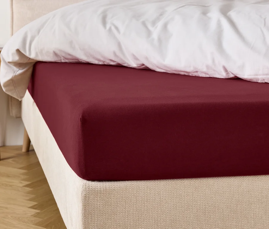 Weinrotes Spannbettlaken auf einem Bett mit weißer Bettdecke.