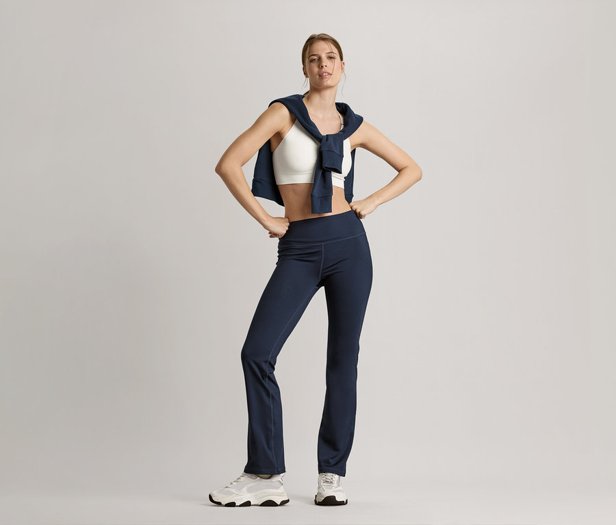 Frau posiert mit Sweatjacke in Navy um die Schultern, weißem Seamless-Sport-Bustier und marineblauer Bodyforming-Sporthose.