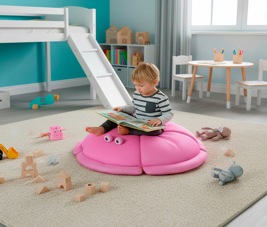 Ein kleiner Junge mit blonden Haaren sitzt auf einem rosafarbenen Krabbenkissen und liest ein Buch. Um ihn herum liegen Holzspielzeuge auf einem Teppich.