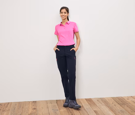 Frau in pinkem Poloshirt und dunkler Hose steht vor einer weißen Wand.