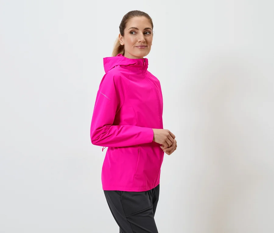 Frau mit Pferdeschwanz trägt pinke Kapuzenjacke und schwarze Hose vor einem weißen Hintergrund.
