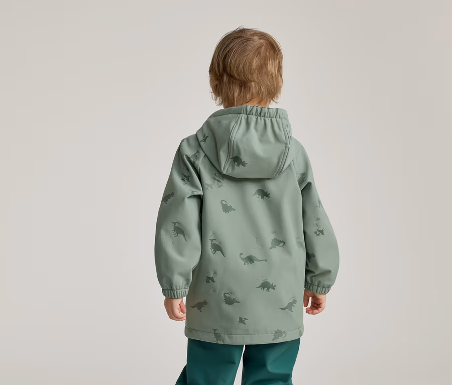Kind von hinten in grüner Dino-Jacke mit Kapuze und grüner Hose vor hellem Hintergrund.