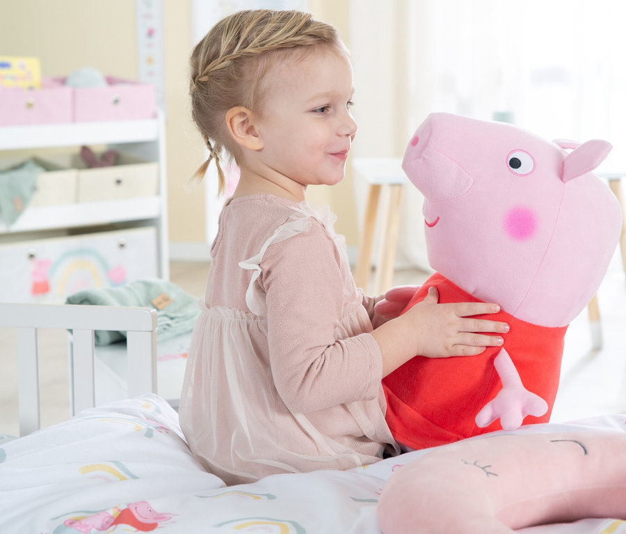 Ein kleines Mädchen sitzt auf einem Bett und umarmt ein Peppa Pig Plüschtier.