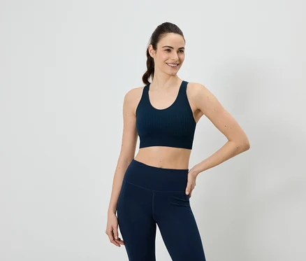 Frau in marineblauem Sport-BH und Leggings, eine Hand an der Hüfte, vor einem weißen Hintergrund.