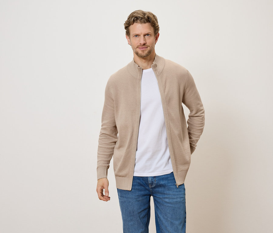 Mann mit hellbraunem Cardigan mit Reißverschluss, weißem T-Shirt und Jeans vor hellem Hintergrund.