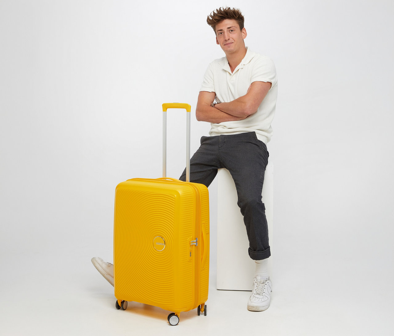 Mann sitzt neben gelbem American Tourister Hartschalen-Koffer »Soundbox« Spinner, groß.
