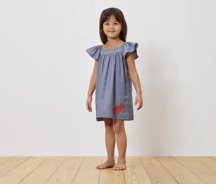 Ein junges Mädchen steht barfuß vor einer weißen Wand und trägt ein blaues Kleid mit Erdbeerapplikationen.