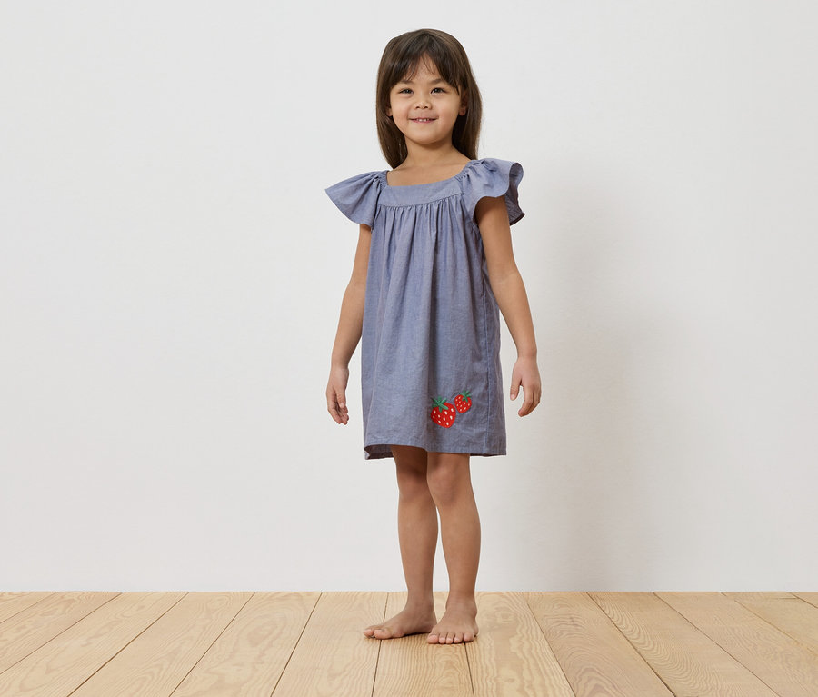 Ein junges Mädchen steht barfuß vor einer weißen Wand und trägt ein blaues Kleid mit Erdbeerapplikationen.