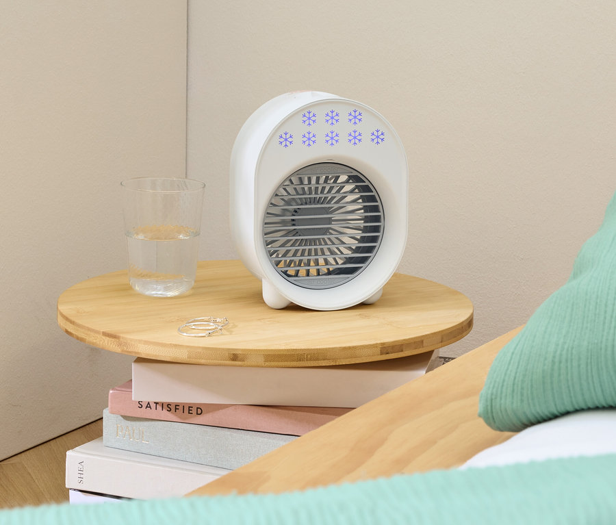 Ein weißer, tragbarer Ventilator mit blauen Schneeflocken steht auf einem kleinen Holztisch neben einem Glas Wasser und Ringen.