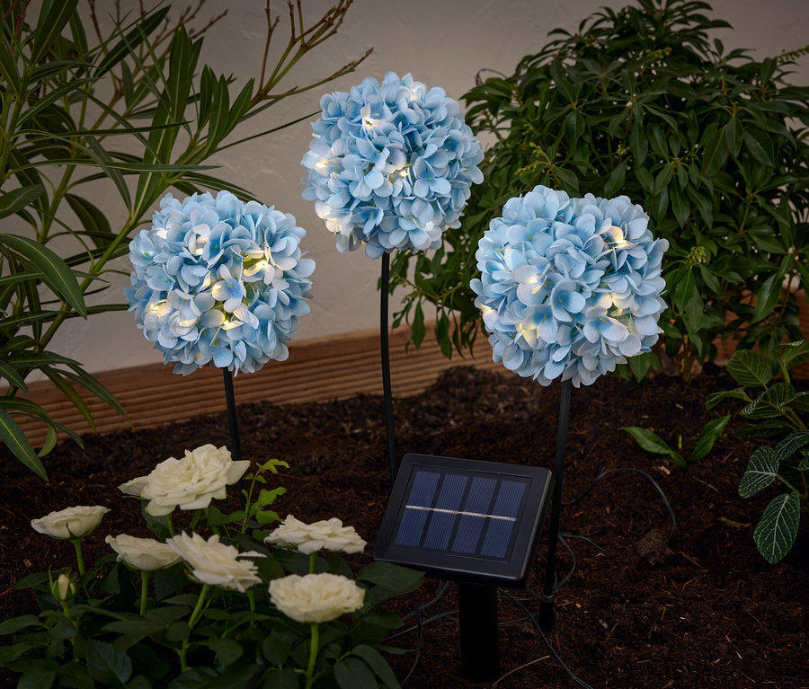 Drei »Hortensie« Solar-Spieße leuchten zwischen weißen Rosen und Pflanzen im Beet.