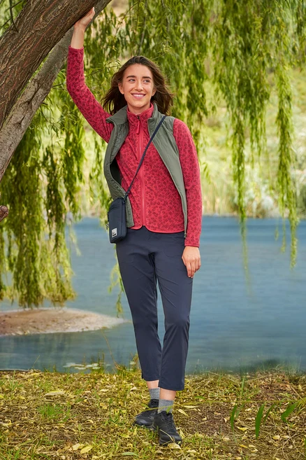 Frau lehnt an Baum am See, trägt rote Fleecejacke, marineblaue Hose und khaki »packable« Hybrid-Weste.