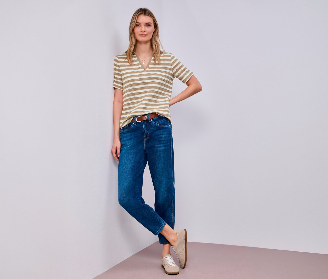Frau posiert mit Street One Strickshirt, Jeans, braunem Gürtel und beigen Schuhen an einer Ecke.