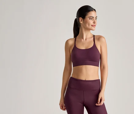 Frau in Sportbekleidung in Pflaumenfarbe, bestehend aus Sport-BH mit dünnen Trägern und Leggings, posiert vor einem hellen Hintergrund.