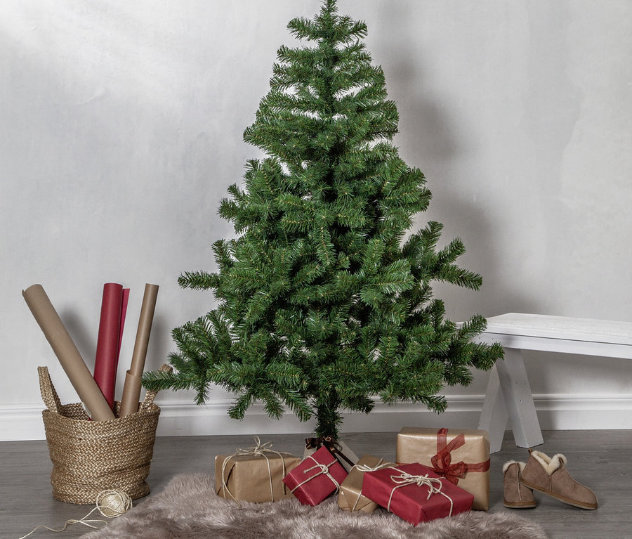 Ein grüner künstlicher Weihnachtsbaum steht auf einem grauen Holzboden, umgeben von braun und rot verpackten Geschenken. Ein hellbrauner Fellteppich liegt unter dem Baum. Links steht ein Korb mit Geschenkpapierrollen und Garn, rechts ein Paar braune Hausschuhe.